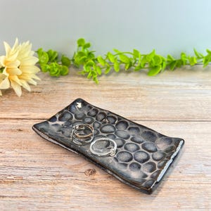 May include: A rectangular ceramic trinket dish featuring a dark gray and black textured surface with a repeating pattern of raised circular indentations. Two metallic rings, one silver and one gold-toned, rest on the center of the dish.