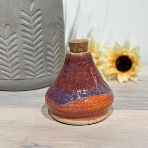 May include: A handmade ceramic bottle with a cork stopper. The bottle features a glossy finish with a color gradient of orange, purple, and red. The base is a light beige color. Decorative item.