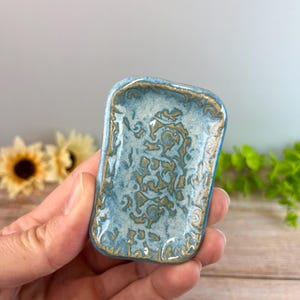 May include: A small rectangular ceramic tray featuring a glossy light blue glaze with a textured, raised gold-toned pattern across the surface. The edges of the tray are slightly rounded and finished with a matching gold-toned decorative trim.