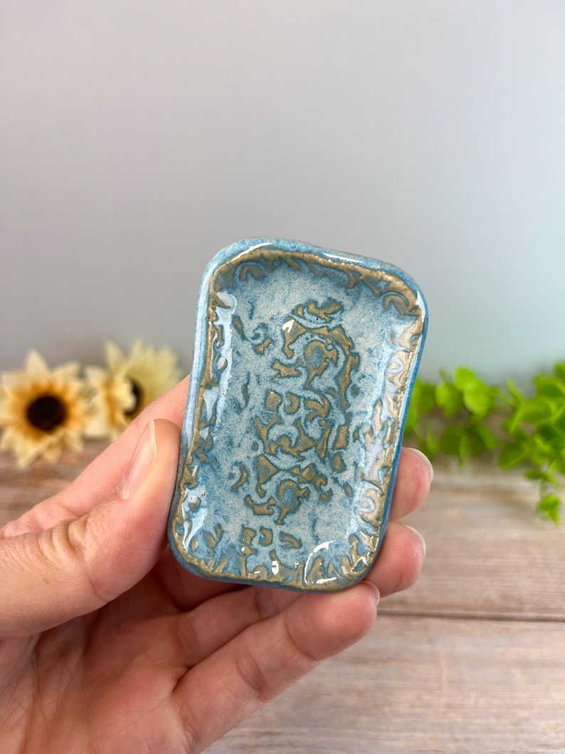 May include: A small rectangular ceramic tray featuring a glossy light blue glaze with a textured, raised gold-toned pattern across the surface. The edges of the tray are slightly rounded and finished with a matching gold-toned decorative trim.