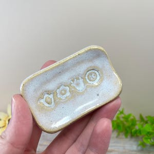 May include: A small, rectangular ceramic dish with a speckled off-white glaze and a yellow-tinted rim. The center of the dish features three embossed, circular floral patterns arranged in a horizontal row, with a smooth, glossy finish.