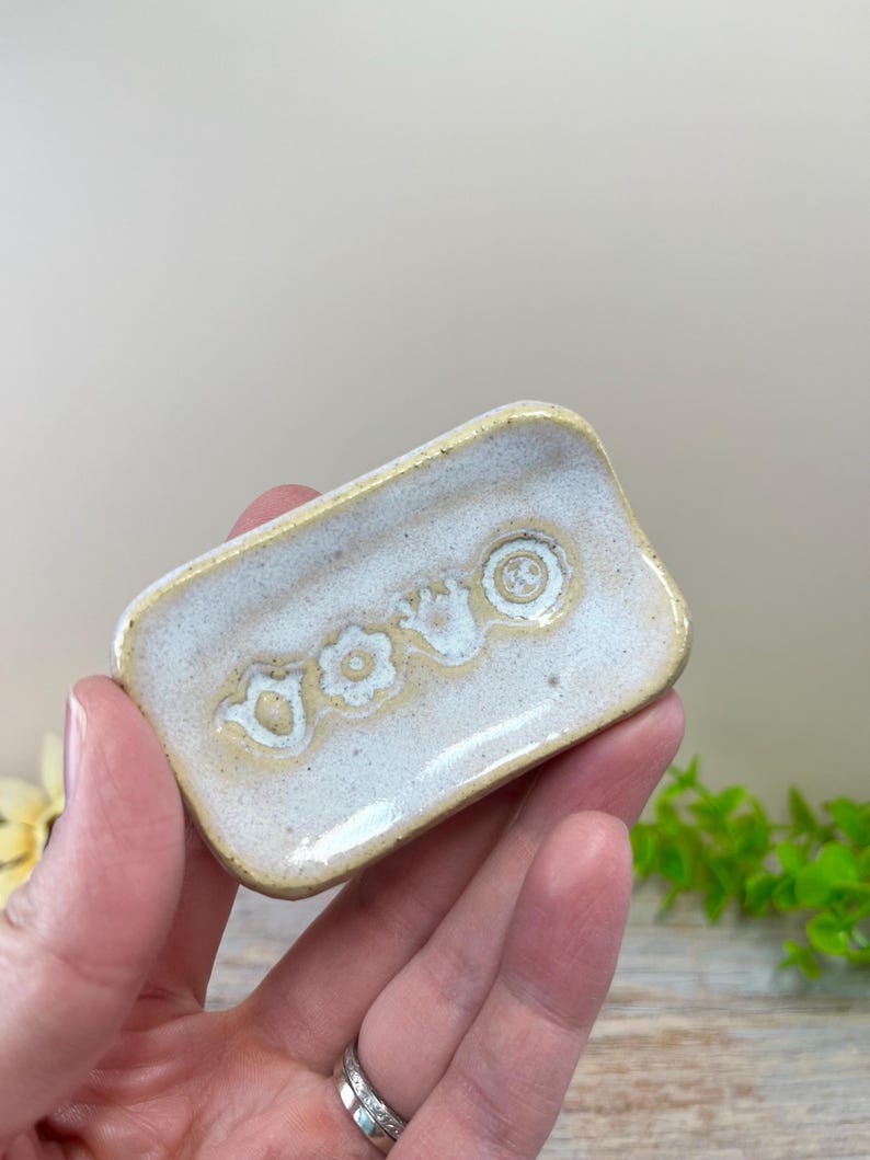 May include: A small, rectangular ceramic dish with a speckled off-white glaze and a yellow-tinted rim. The center of the dish features three embossed, circular floral patterns arranged in a horizontal row, with a smooth, glossy finish.