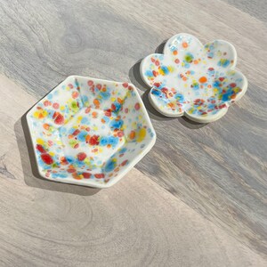 May include: Two small, white ceramic dishes with colorful speckled designs. One is a hexagonal shape, and the other is a flower shape. The speckled patterns include red, blue, orange, and yellow dots. These decorative dishes are ideal for holding small items.