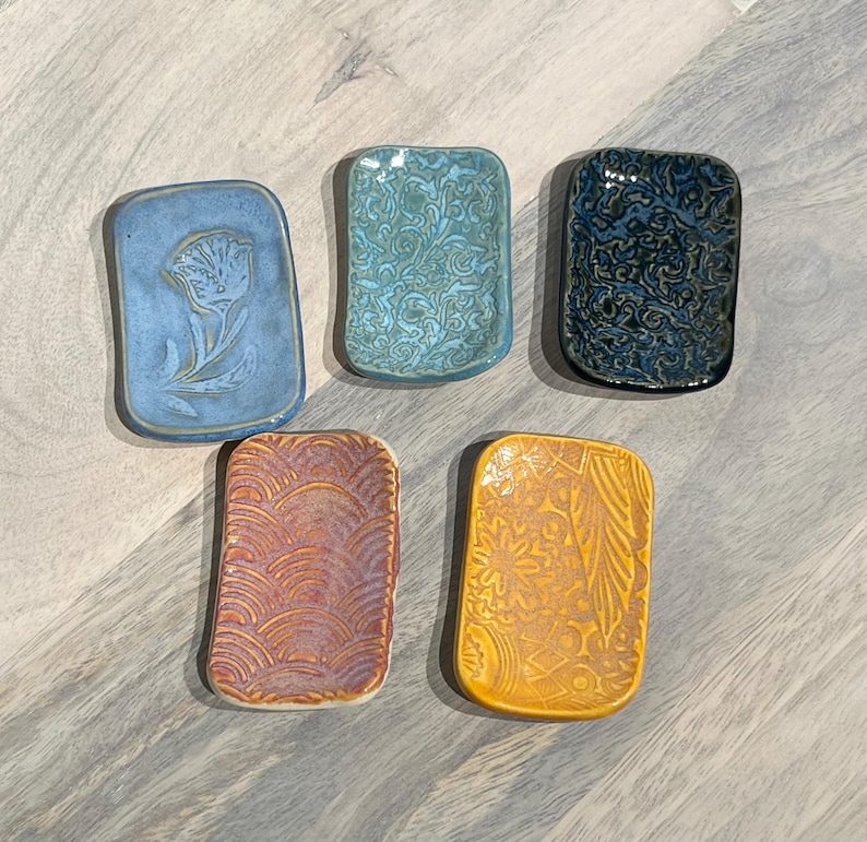 May include: Five rectangular ceramic trinket dishes with rounded corners. The top row features a light blue dish with a flower, a teal dish with a swirling pattern, and a dark blue dish with a textured design. The bottom row has a purple dish with a wave pattern and a yellow dish with a floral motif.