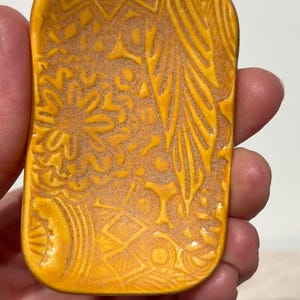 May include: A rectangular ceramic dish with rounded corners, featuring a vibrant mustard yellow glaze. The surface is decorated with an intricate, embossed pattern of geometric shapes, floral motifs, and linear textures across the entire face.
