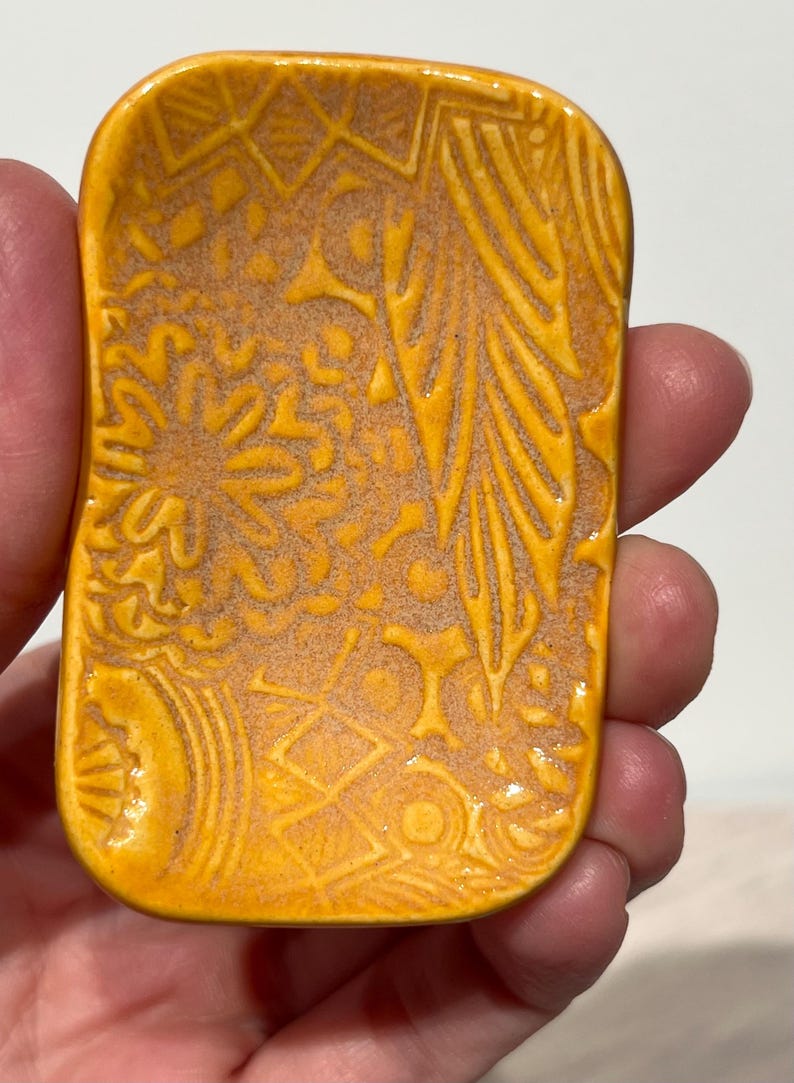 May include: A rectangular ceramic dish with rounded corners, featuring a vibrant mustard yellow glaze. The surface is decorated with an intricate, embossed pattern of geometric shapes, floral motifs, and linear textures across the entire face.