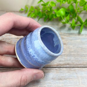 May include: A small, handmade ceramic cup with a light blue glaze. The cup has a rounded shape with a textured, ridged exterior and a white base. The interior is a darker shade of blue. The cup is held in a hand.