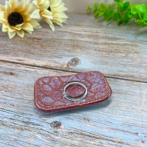 May include: A rectangular ceramic trinket dish with a textured, reddish-brown glaze featuring a repeating lattice pattern. A simple silver-toned metal ring rests in the center of the dish, which has slightly raised, rounded edges and a glossy finish.