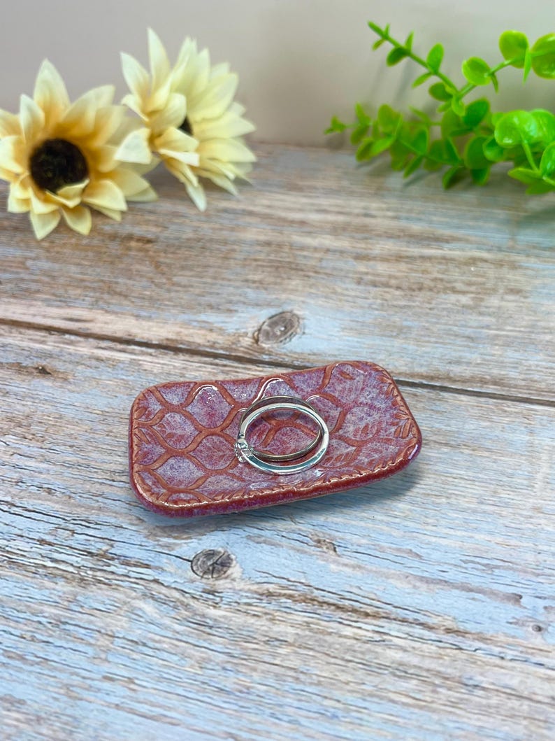 May include: A rectangular ceramic trinket dish with a textured, reddish-brown glaze featuring a repeating lattice pattern. A simple silver-toned metal ring rests in the center of the dish, which has slightly raised, rounded edges and a glossy finish.