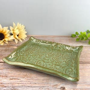May include: A rectangular ceramic tray with a glossy sage green glaze and a raised, intricate swirling scrollwork pattern across the surface. The tray features slightly upturned edges and a smooth, reflective finish throughout the entire piece.