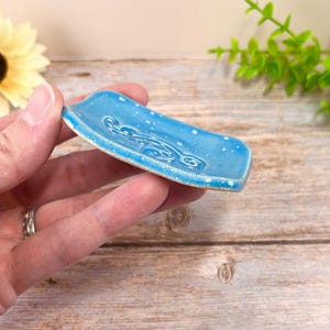 May include: A small, rectangular ceramic dish with a vibrant blue glaze and a speckled white finish. The surface features a raised, swirling embossed pattern in the center, and the edges of the dish are slightly curved upward for a shallow basin.