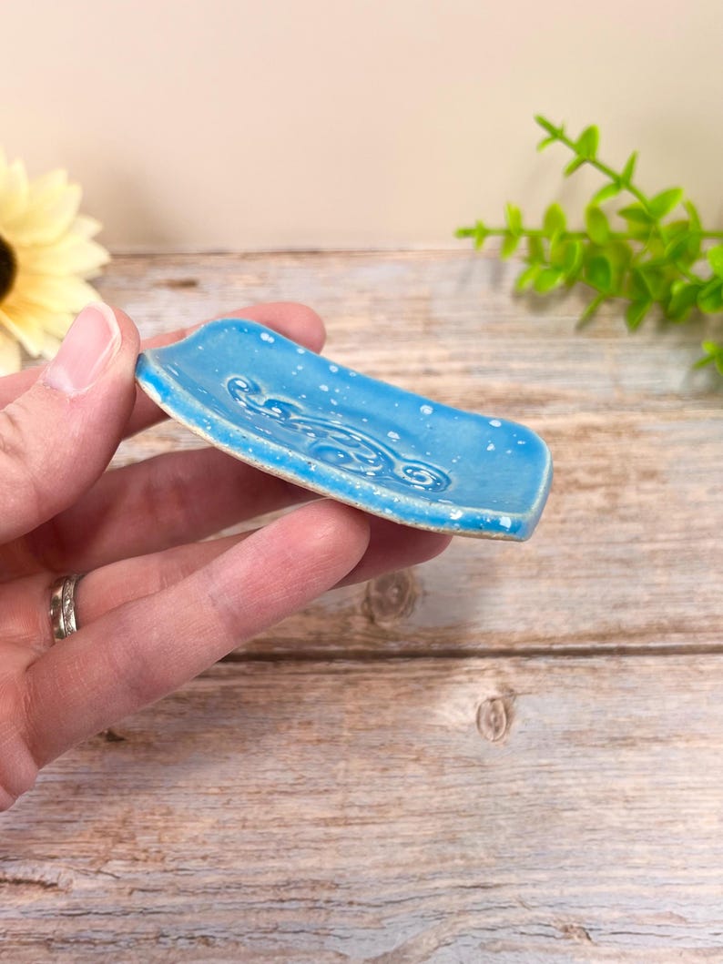 May include: A small, rectangular ceramic dish with a vibrant blue glaze and a speckled white finish. The surface features a raised, swirling embossed pattern in the center, and the edges of the dish are slightly curved upward for a shallow basin.