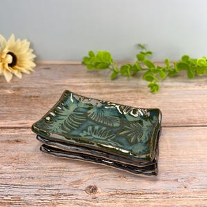 May include: A stack of two rectangular ceramic dishes featuring a glossy dark green glaze with a textured leaf pattern. The edges of the dishes are slightly raised and have a dark, uneven finish, showcasing a handcrafted appearance.