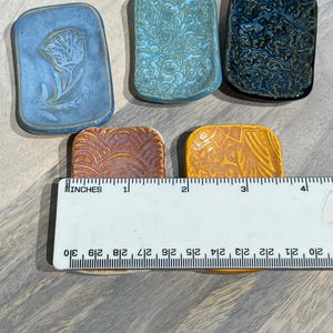 May include: Five rectangular ceramic dishes with varied glazes and textures, including blue, teal, dark blue, rust, and yellow. A white ruler is placed across the bottom two dishes, showing each dish is approximately 2 inches wide in total.