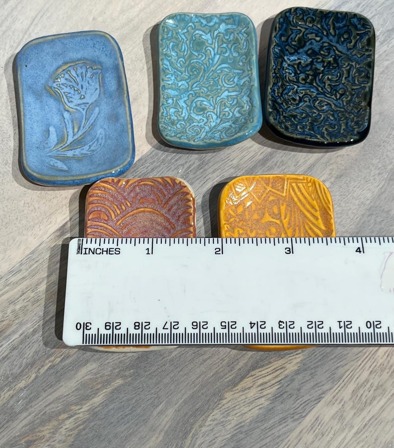 May include: Five rectangular ceramic dishes with varied glazes and textures, including blue, teal, dark blue, rust, and yellow. A white ruler is placed across the bottom two dishes, showing each dish is approximately 2 inches wide in total.
