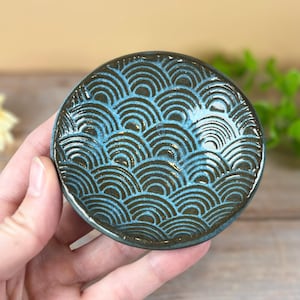May include: A small, round ceramic dish with a wave pattern in shades of blue and brown. The dish is held in a hand, showcasing the detailed design and glossy finish. The wave pattern is a repeating design.