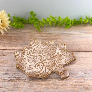 May include: A ceramic trinket dish shaped like a maple leaf, featuring a white glazed surface with intricate, embossed golden-brown swirling patterns and a matching golden-brown rim, designed with a shallow, concave center for holding small items.