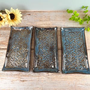 May include: Three rectangular ceramic trays featuring a glossy blue glaze over a dark brown base. Each tray displays an intricate, embossed pattern of swirling vines, leaves, and geometric textures across the entire surface with raised edges.