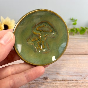May include: A small, round, ceramic dish with a glossy olive green glaze. The center of the dish features a debossed, stylized mushroom design with golden-brown accents that contrast against the smooth, reflective surface of the pottery.