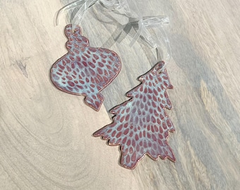 May include: Two ceramic Christmas ornaments. One is an oval shape, and the other is a Christmas tree shape. Both ornaments have a maroon and blue speckled pattern and are strung with sheer white ribbon for hanging.