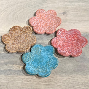 May include: Four ceramic flower-shaped dishes in shades of pink, blue, and tan. Each dish features a detailed, embossed pattern. The dishes are arranged on a light wood-grain surface.
