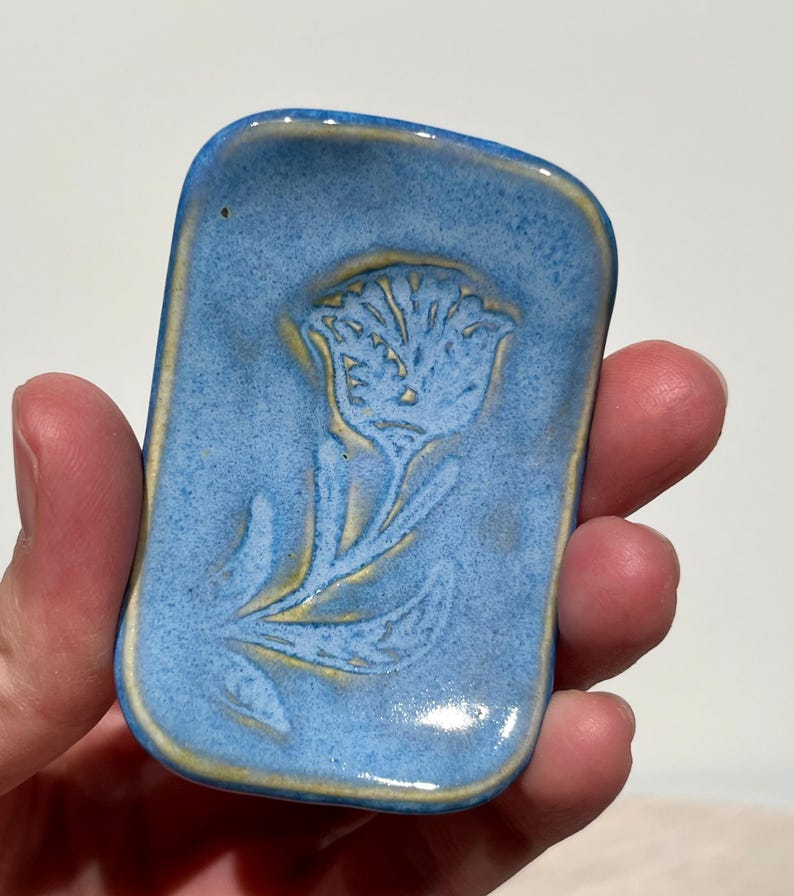 May include: A rectangular ceramic dish with rounded corners, featuring a light blue speckled glaze and a debossed floral design in the center. The edges and the recessed flower pattern reveal a warm, yellowish-tan clay base beneath the blue glaze.