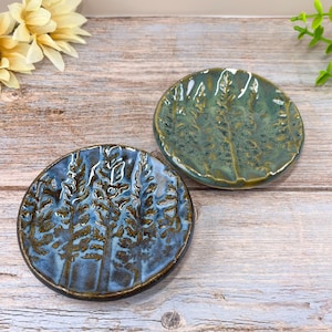 May include: Two handmade ceramic dishes with a forest design. One dish is blue with brown accents, and the other is teal. The dishes are round and have a textured surface, perfect for holding small items.