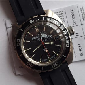 Op de afbeelding: Een zwarte Vostok duikershorloge met een zwarte bezel en een zwarte rubberen band. De wijzerplaat heeft een duikergrafiek, lichtgevende markeringen en de tekst "Восток". De horloge heeft een roestvrijstalen kast en een datumvenster.