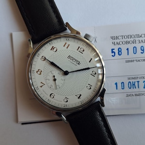 May include: A classic wristwatch with a white dial, black hands, and a black leather strap. The watch face displays Arabic numerals and a subdial. The watch is marked "VOSTOK 17 KAMHEЙ" and is set against a white background.