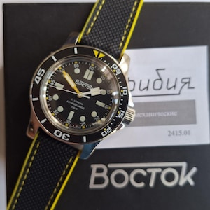 May include: A black and silver wristwatch with a black dial, white markings, and yellow hands. The watch has a black strap with yellow stitching. The watch face reads "Восток" and "200 M".