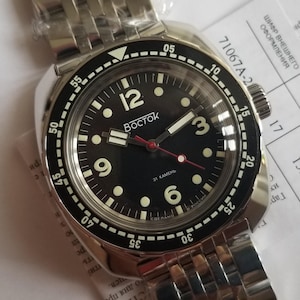 Könnte beinhalten: Eine Vostok-Uhr aus Edelstahl mit schwarzem Zifferblatt. Die Uhr hat weiße Ziffern, Leuchtzeiger und einen roten Sekundenzeiger. Die Lünette hat ein schwarz-weißes Design. Das Armband ist aus Edelstahl.