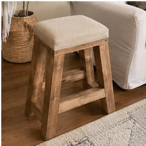 Puede incluir: Un taburete cuadrado con estructura de madera natural en tono marrón medio y cuatro patas robustas conectadas por barras de soporte horizontales. La parte superior del taburete está tapizada en tela beige claro, ofreciendo una superficie de asiento plana.