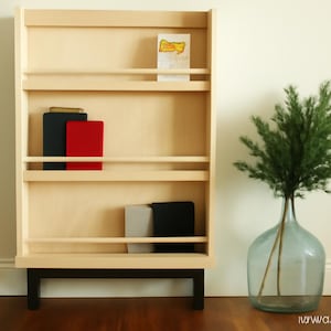 Op de afbeelding: Een licht houten boekenplank met zwarte poten, met boeken en een wenskaart. De planken hebben een eenvoudig ontwerp. Een heldere glazen vaas met een groene plant staat in de buurt. De kamer heeft een minimalistische uitstraling.