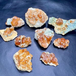 Pode incluir: Uma coleção de pedras preciosas brutas, não cortadas, em tons de laranja e castanho. As pedras preciosas estão agrupadas numa superfície escura, mostrando as suas texturas naturais e estruturas cristalinas. Algumas estão incrustadas numa matriz rochosa.