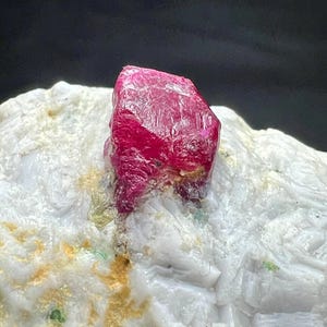 Puede incluir: Una vibrante gema de rubí facetada, que muestra un tono rojo intenso, se encuentra sobre un fondo de roca blanca y amarilla. La estructura cristalina del rubí es visible, destacando su belleza natural. El espécimen mineral es un ejemplo llamativo de geología natural.