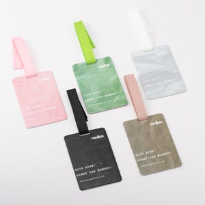 May include: A collection of rectangular luggage tags in various colors: pink, green, black, gray, and tan. Each tag has a matching ribbon loop and the text "relax" and "SLOW DOWN + SCENT THE MOMENT +" printed on it.