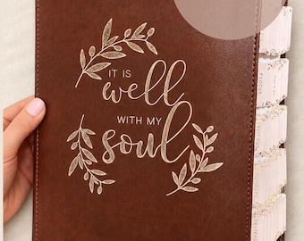 May include: A brown leather-like Bible cover featuring the engraved text 'IT IS well WITH MY soul' surrounded by olive branch illustrations. The name 'Debra' is engraved at the bottom, and the side displays white index tabs for book sections.