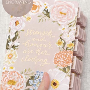 Peut inclure: Un journal à thème floral avec le texte "Strength and honour are her clothing." La couverture est rose blush, ornée de fleurs peintes en pêche, blanc et bleu. Le journal a des onglets étiquetés et le nom "Emily" est imprimé en bas à droite.