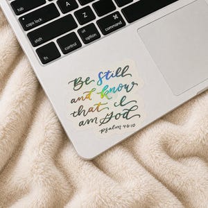 May include: A holographic sticker featuring the cursive text Be still and know that I am God and the reference Psalm 46:10. The sticker has a multi-colored iridescent finish that shifts in appearance, applied to the surface of a silver laptop.