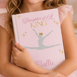 Op de afbeelding: Een roze boek met de titel "Daughter of A KING", een ballerina-afbeelding en de naam "Callie". De kaft heeft gouden tekst en steraccenten. Het boek wordt door een persoon vastgehouden.