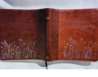 Könnte beinhalten: Eine braune Leder-Bibel mit umlaufendem Einband. Vorder- und Rückseite zeigen geprägte Wildblumen-Illustrationen in Metallic-Farben. Der Buchrücken trägt den Text "Journaling Study Bible" und vorne steht "Consider how the wildflowers grow".