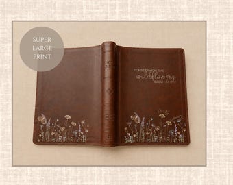 Könnte beinhalten: Ein brauner Bibeleinband aus Leder mit einer Illustration von Wildblumen und Schmetterlingen am unteren Rand, sowie dem Text "CONSIDER HOW THE wildflowers GROW" und "SUPER LARGE PRINT" auf dem Einband und einem runden Overlay.