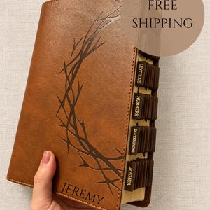 Puede incluir: Una cubierta de libro de cuero sintético marrón con un diseño de corona de espinas grabado con láser y el nombre JEREMY. El libro incluye pestañas de índice de color marrón oscuro etiquetadas con nombres de libros bíblicos como EXODUS, LEVITICUS, NUMBERS, DEUTERONOMY y JOSHUA.