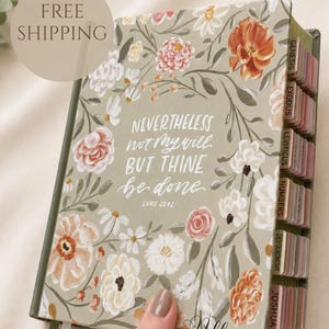 May include: A sage green hardcover Bible featuring a floral pattern with white, pink, and orange flowers. The cover displays the white text "NEVERTHELESS not My will BUT THINE be done LUKE 22:42" and the name "Allison" in black script. Includes index tabs.