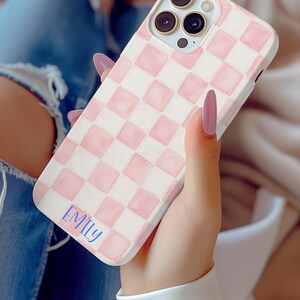 May include: A phone case with a pink and white checkered pattern. The case has a soft, watercolor-like appearance. The name "EMILY" is written in blue script on the lower left corner of the case.