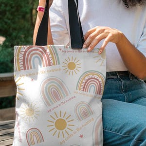 Op de afbeelding: Witte draagtas met een herhalend patroon van pastelkleurige regenbogen, zonnen en sterren. De tekst "Joy comes in the morning" is op het ontwerp gedrukt. De tas heeft zwarte handvatten.