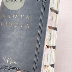 May include: Close-up of a gray leather Bible with gold lettering that reads "SANTA BIBLIA" and "Silvia". The book has decorative corner accents and tabbed pages. The text "FREE ENGRAVING" is also visible.