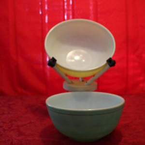 May include: Two vintage mixing bowls, one light green and one off-white, are displayed. The off-white bowl is held by a yellow and wooden stand. The bowls are set against a red backdrop.