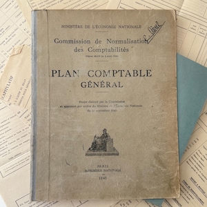 May include: A vintage French book, "PLAN COMPTABLE GENERAL", published in 1947. The cover is a faded tan colour with black text and a small illustration. Other aged documents surround the book.