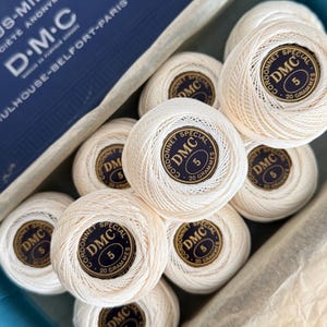 Könnte beinhalten: Eine Sammlung von cremefarbenen DMC Cordonnet Special Baumwollgarnknäueln, jeweils mit der Aufschrift "5" und "20 GRAMMES". Die Verpackung enthält eine blaue Schachtel mit dem DMC-Logo und Text. Geeignet für Häkel- und Stickprojekte.